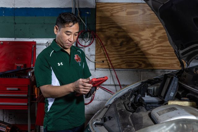 Mechanic in a green shirt holding jumper cables, looking at a car engine. Shop setting.