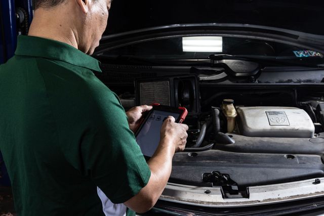 Mechanic using a diagnostic tablet in a car engine bay, wearing a green shirt.