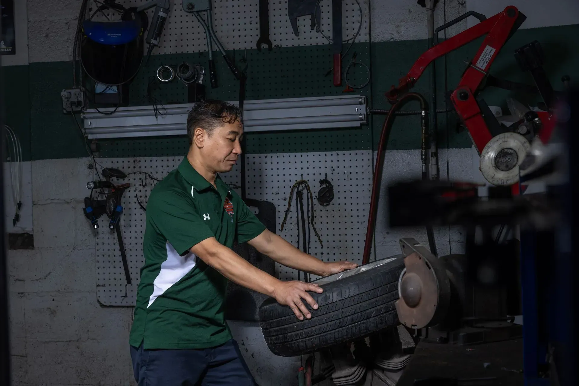 Mechanic in green shirt holding a tire in a workshop with tools in the background.