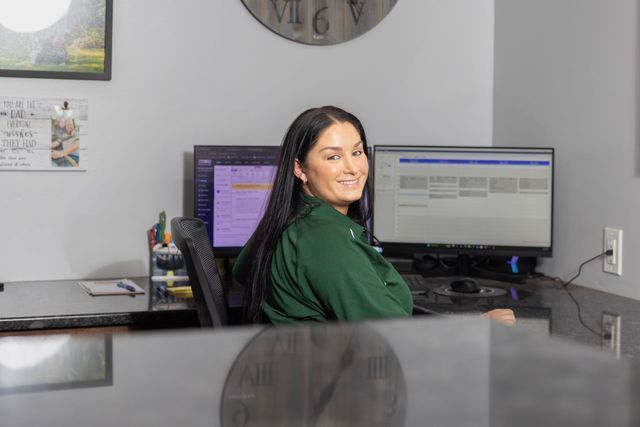 Woman sitting at a desk in an office smiles towards the camera, with two computer monitors behind her.