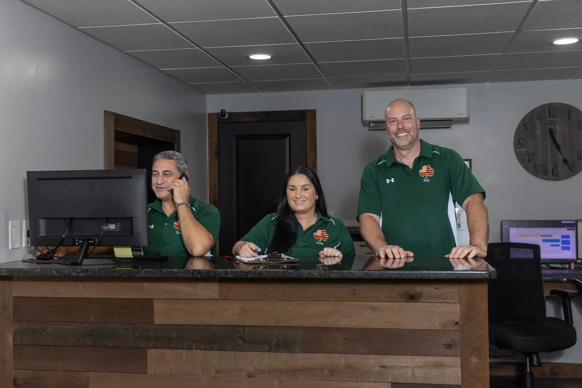 Three people at a reception desk in green shirts. The woman on the left is on the phone. The other two are smiling.