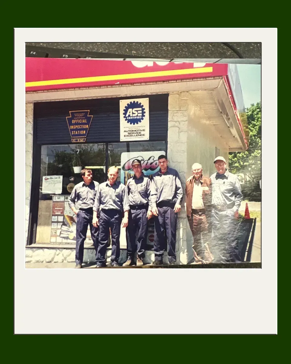 Group of six men standing in front of a gas station. Men are in work uniforms and casual wear. Red and yellow awning.