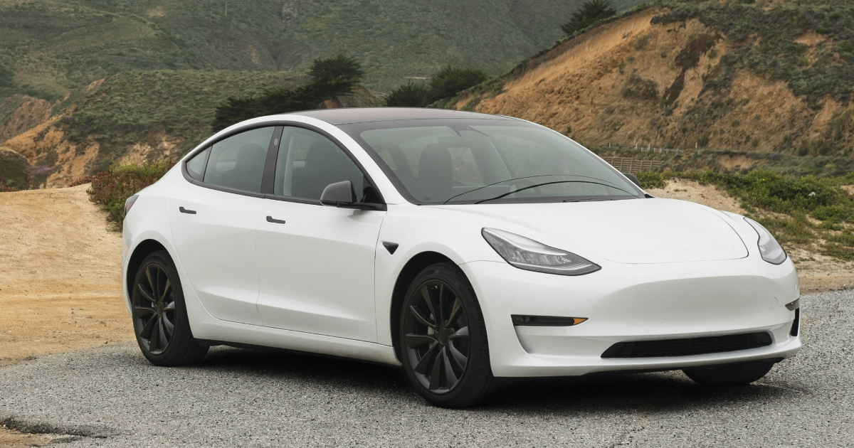 White Tesla Model 3 car parked on gravel road with hillside background.