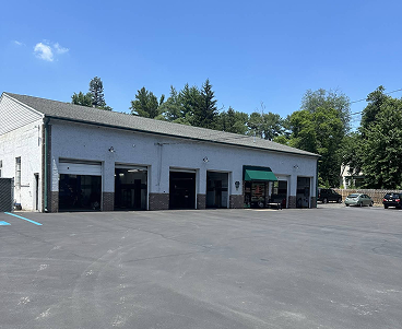 Exterior of a gray building with multiple garage bays, set against a blue sky, with some vehicles in the parking lot.