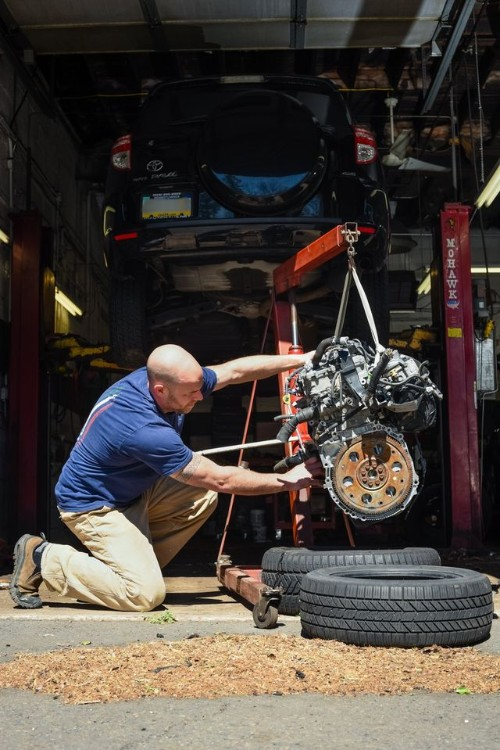 Mechanic kneeling, installing a vehicle engine in a garage with a car lifted on a hoist.