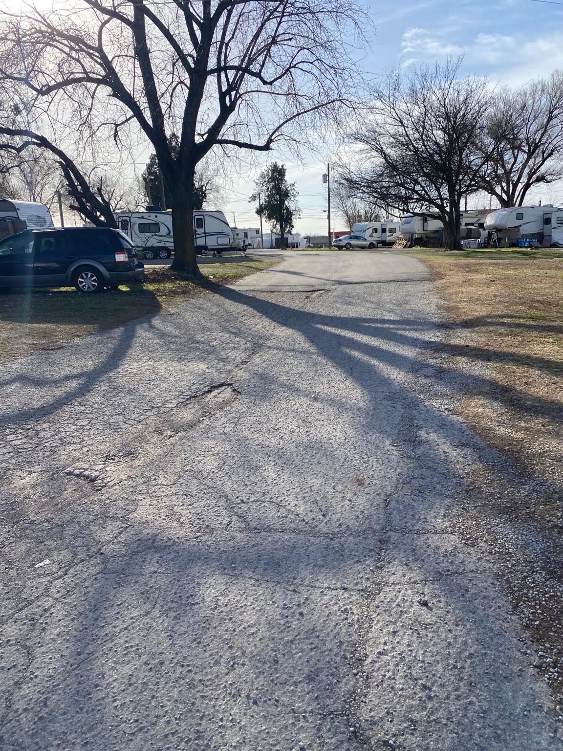 A gravel road leads through a campground with parked RVs and trees casting long shadows on a sunny day.