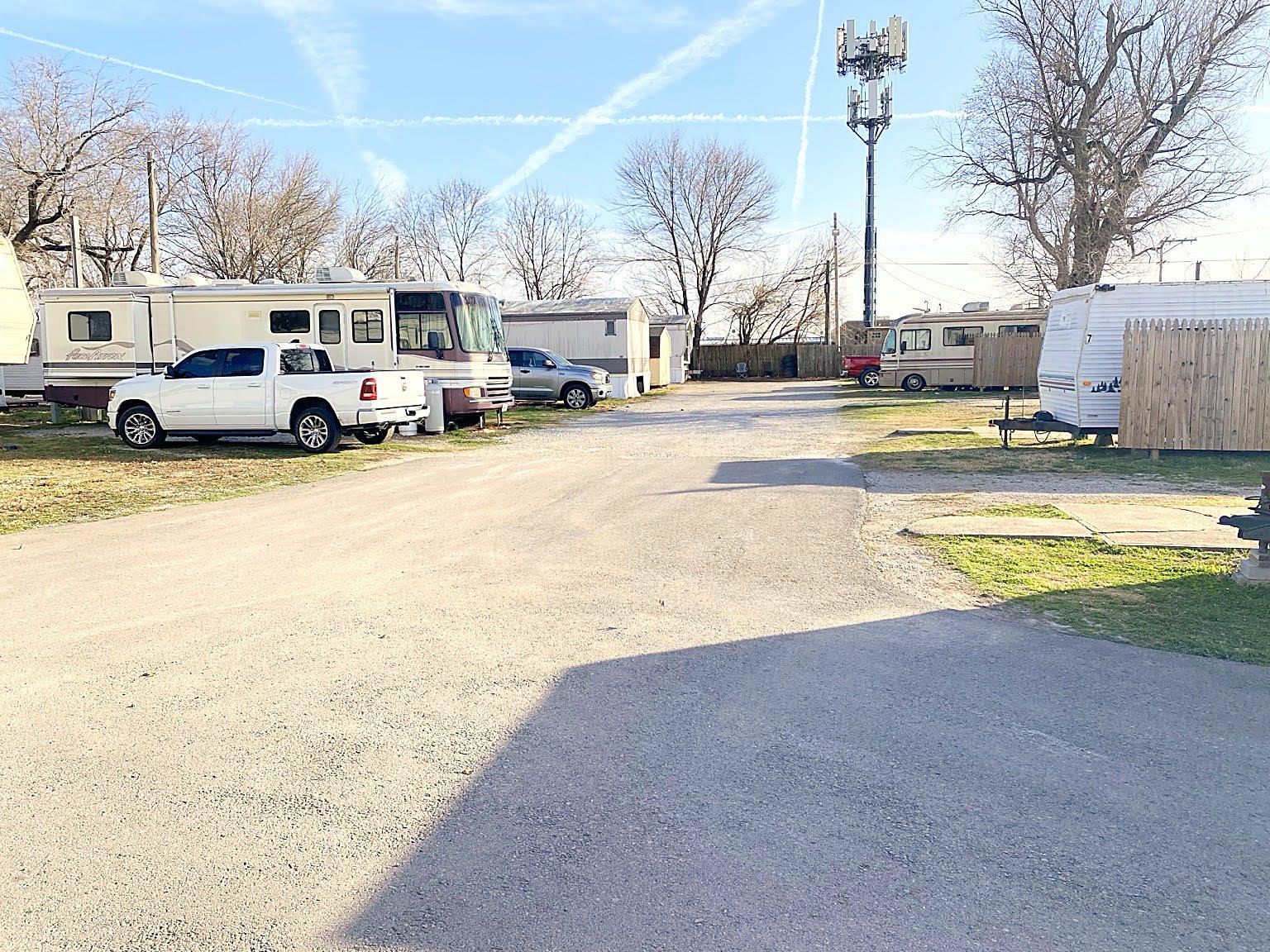 A gravel lot featuring several parked RVs, a white pickup truck, and a tall cell tower under a clear blue sky.