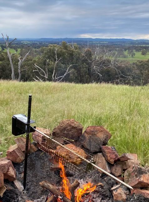 A rotisserie spit roasts meat over a campfire on a grassy hill overlooking a scenic, tree-covered valley.