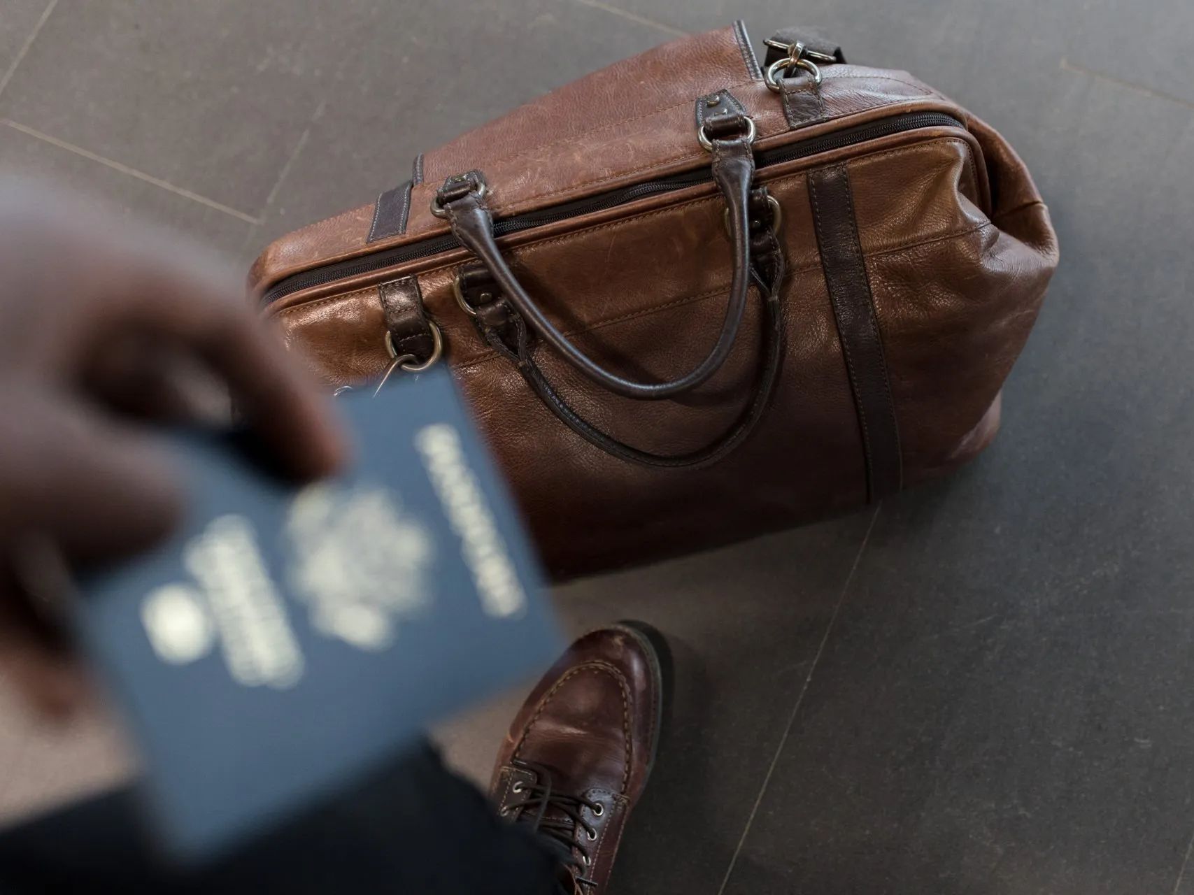 Une personne tenant un passeport près d'un sac de voyage en cuir marron, sur un sol sombre.