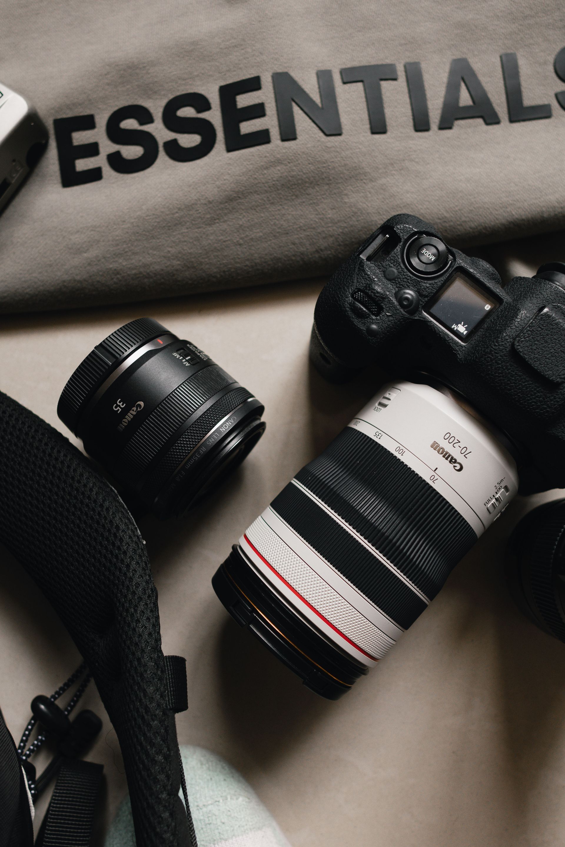 Matériel photo comprenant un reflex numérique, un objectif et un sac gris « ESSENTIALS » sur une surface sombre