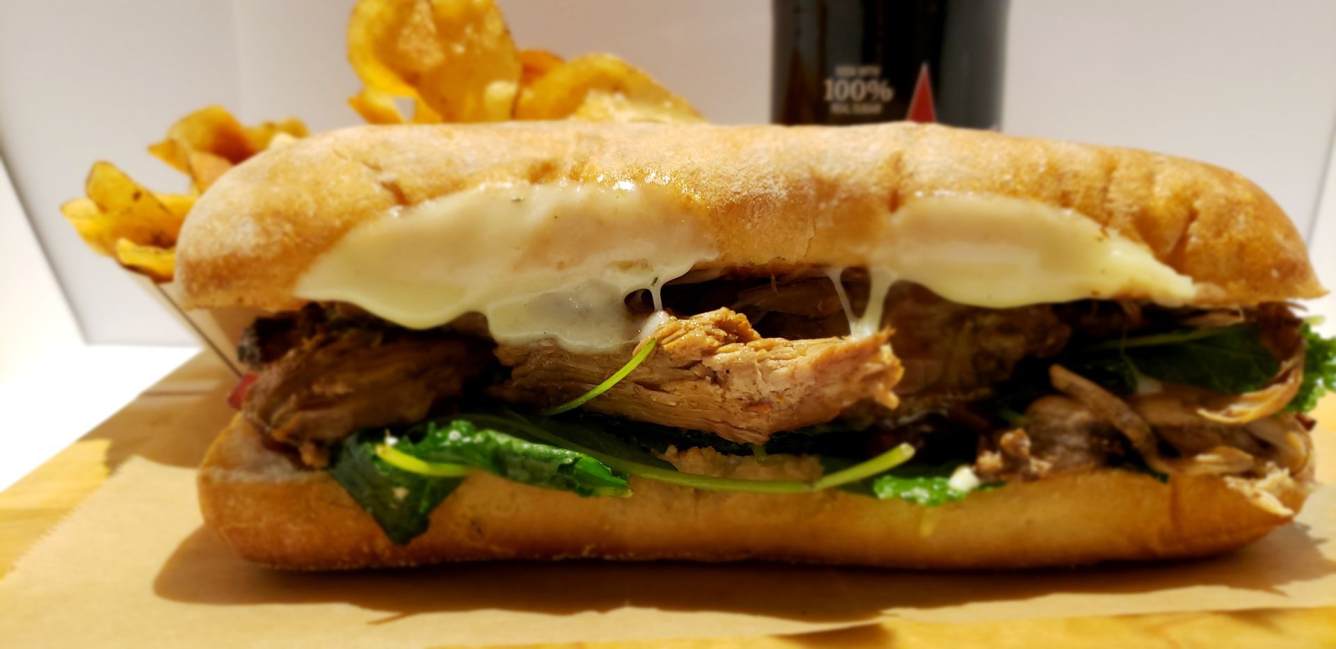 A deli sandwich with melted cheese, greens, and meat. Side of fries and a dark drink in the background.