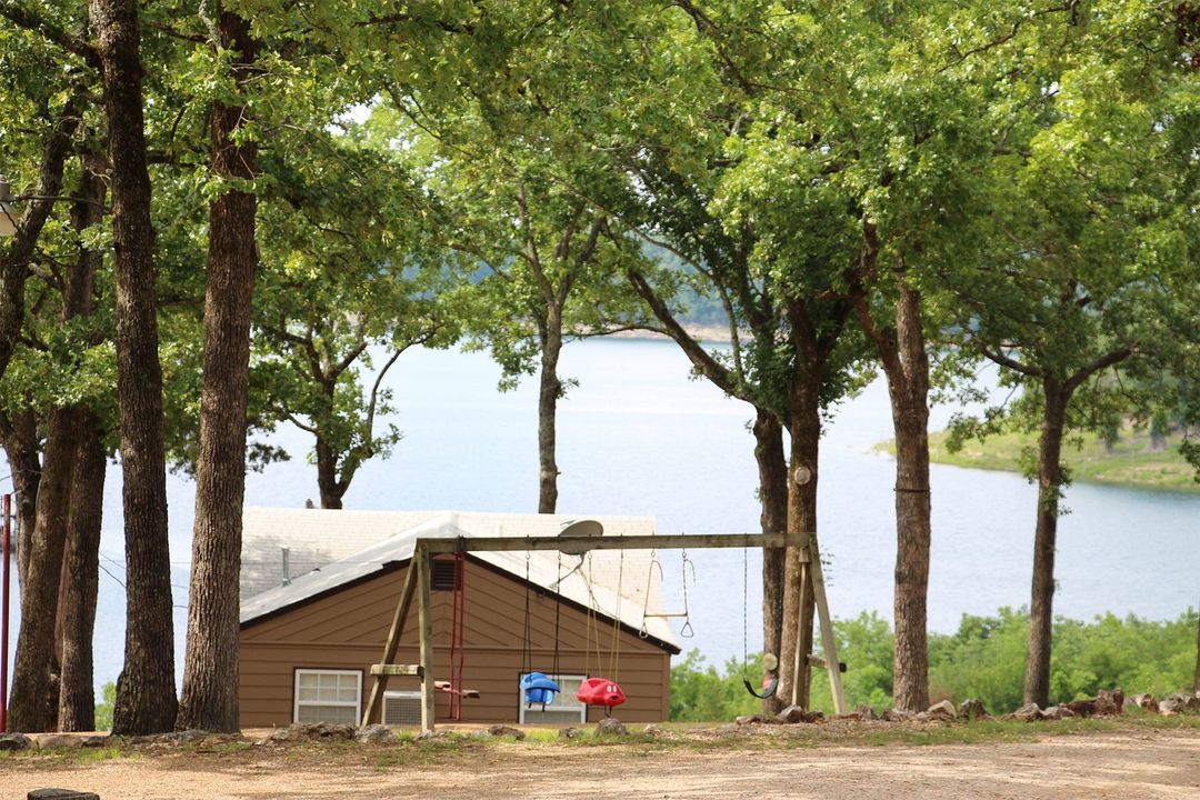 A house with a swing set in front of a lake surrounded by trees.