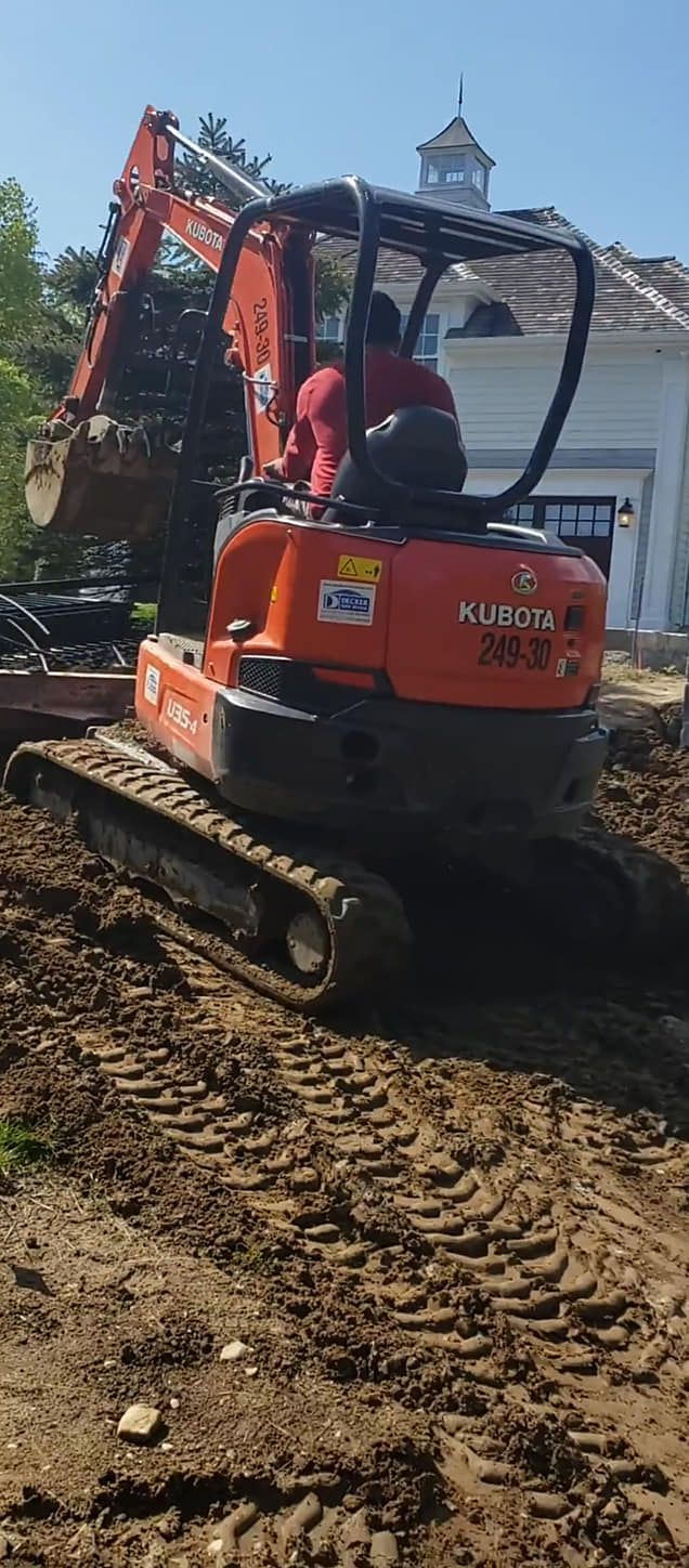 An orange excavator digs in a muddy construction site near a white house. A worker operates the machine.