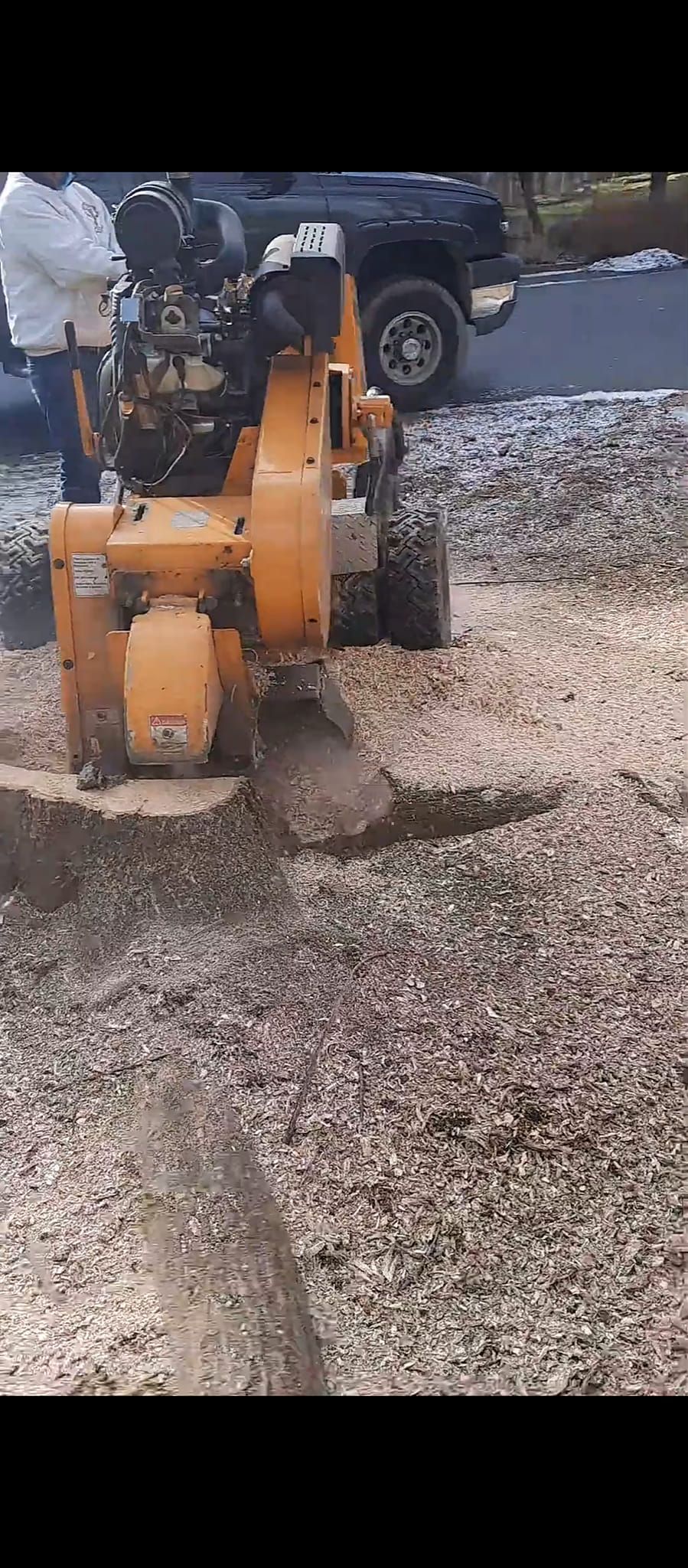 A machine grinding a trench in gravel. A person stands nearby. A vehicle is in the background.