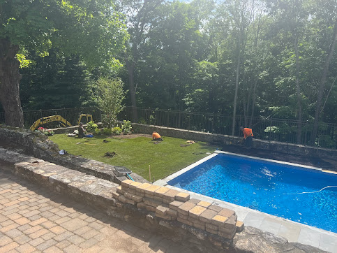 A pool surrounded by a stone patio and retaining wall. Two workers in orange vests are near the new grass area. 