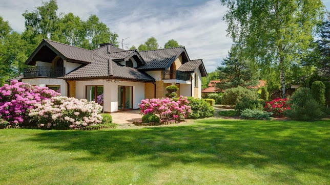 Two-story house with green lawn, pink flowering bushes, and trees under a sunny sky.