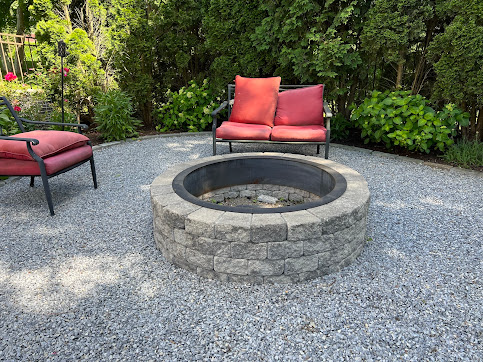 Stone fire pit with seating on a gravel patio in a garden setting.