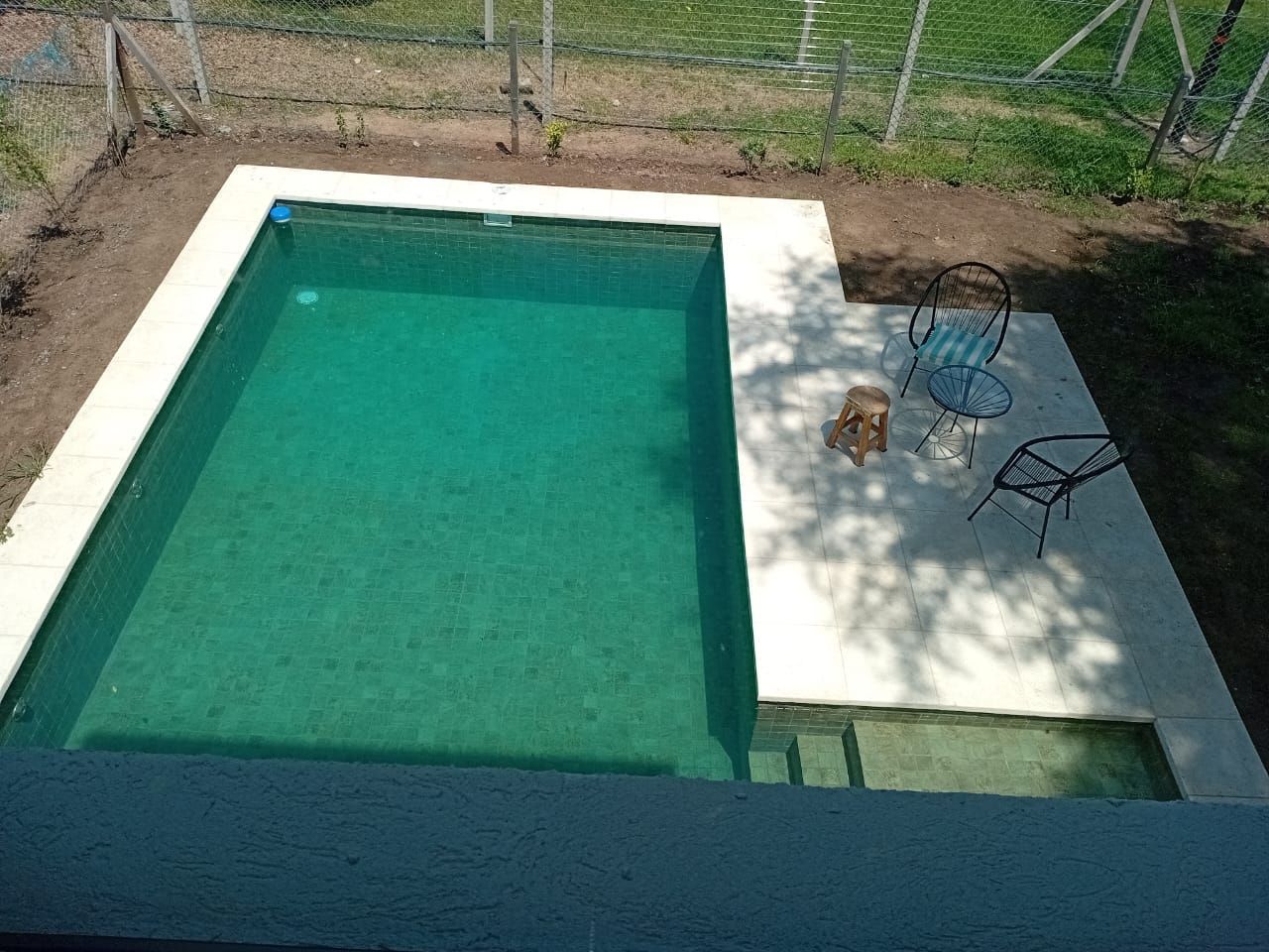 Piscina rectangular de color turquesa con borde de concreto blanco, área de patio pequeño, dos sillas.