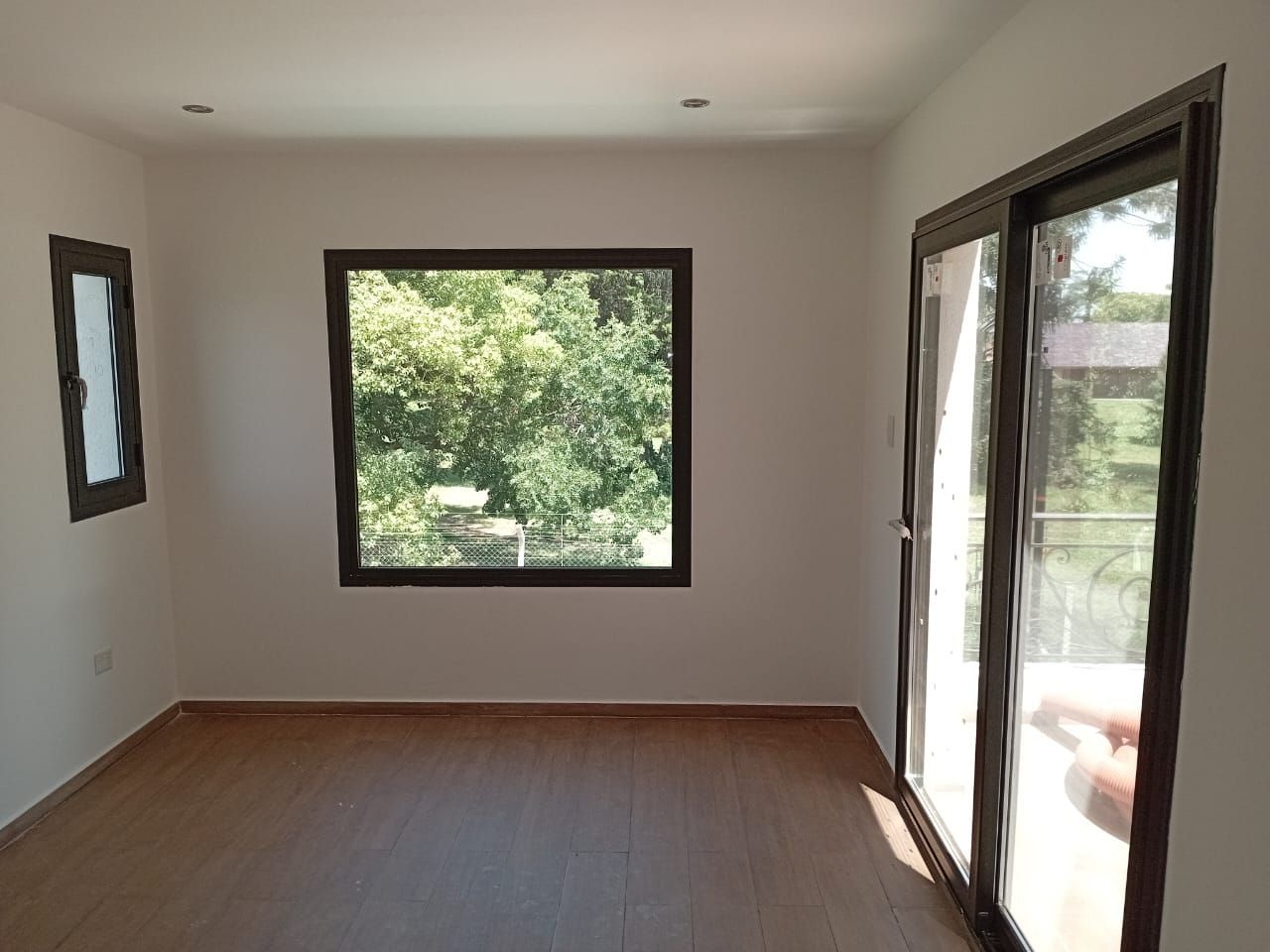 Habitación vacía con tres ventanas de marco oscuro; piso de madera y paredes blancas.