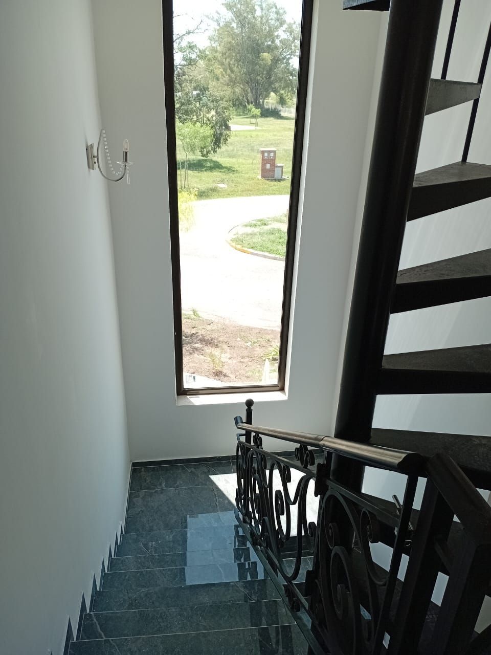 Escalera con barandilla oscura y ventana estrecha con vista a un paisaje verde.