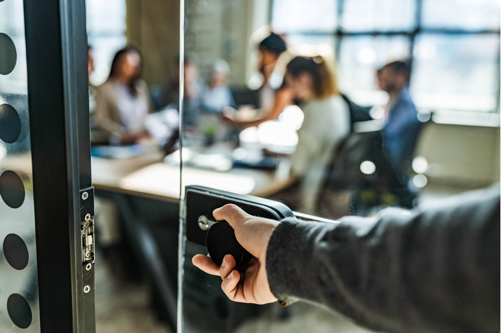 A person is opening a glass door in an office with a group of people in the background.