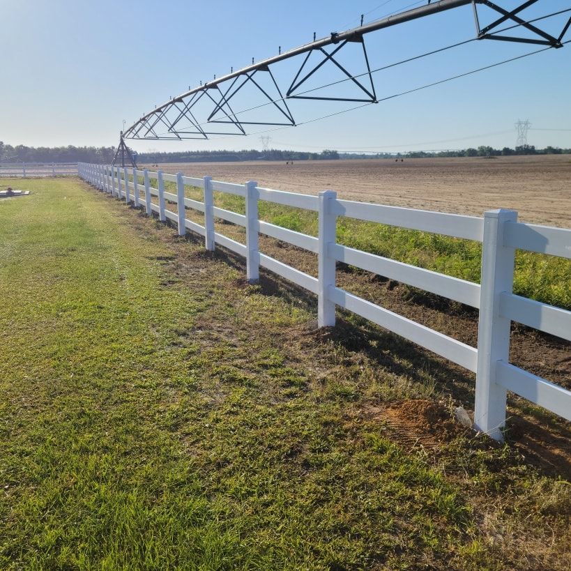 A white fence surrounds a grassy field with a irrigation system in the background.
