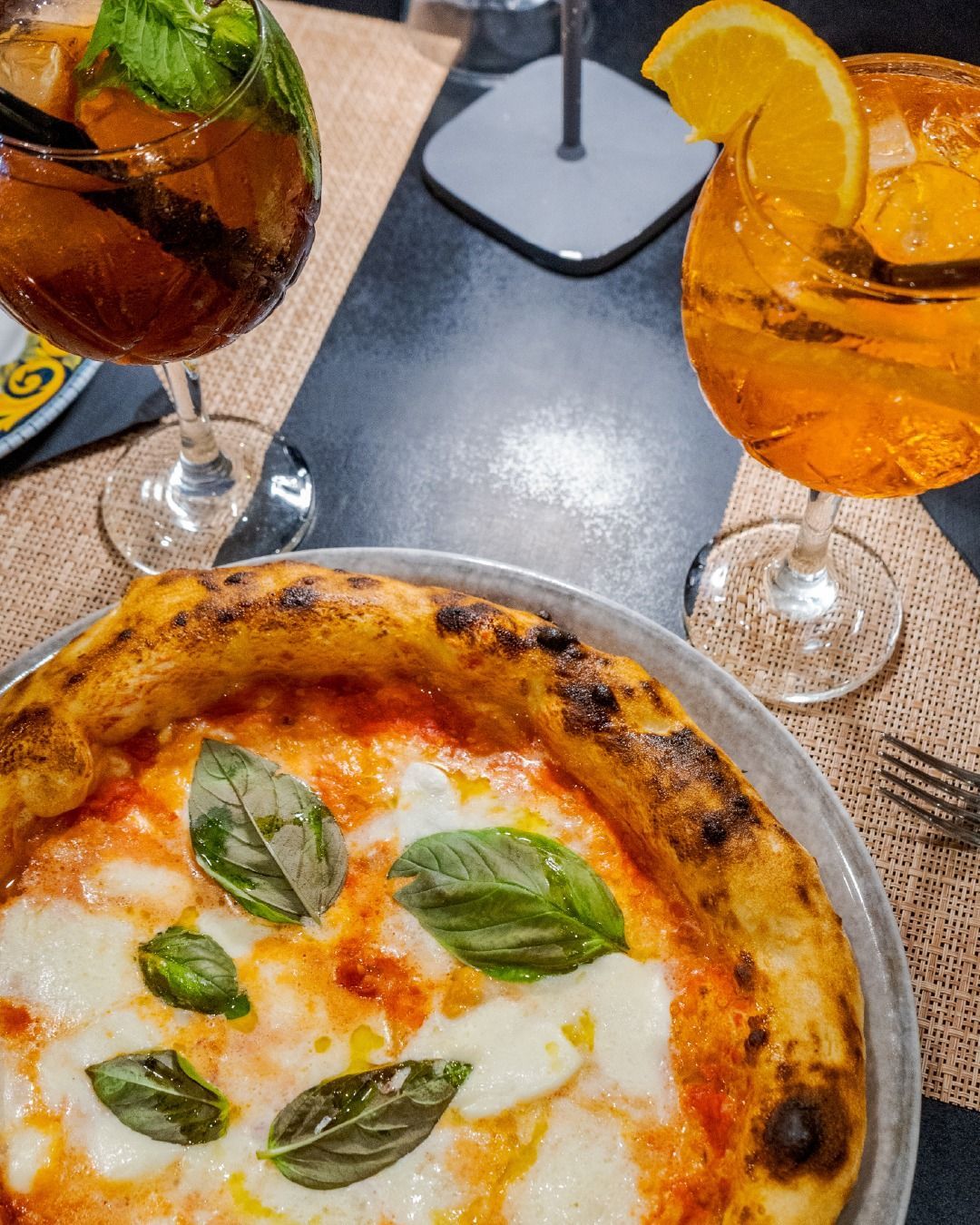 Primo piano di una pizza condita con basilico fresco, accompagnata da due cocktail in calici di vetro su un tavolo di un ristorante.