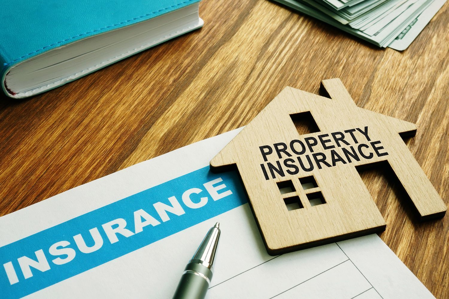 Property insurance documents with house-shaped cutout and pen on a wooden table, with money and a book in the background.