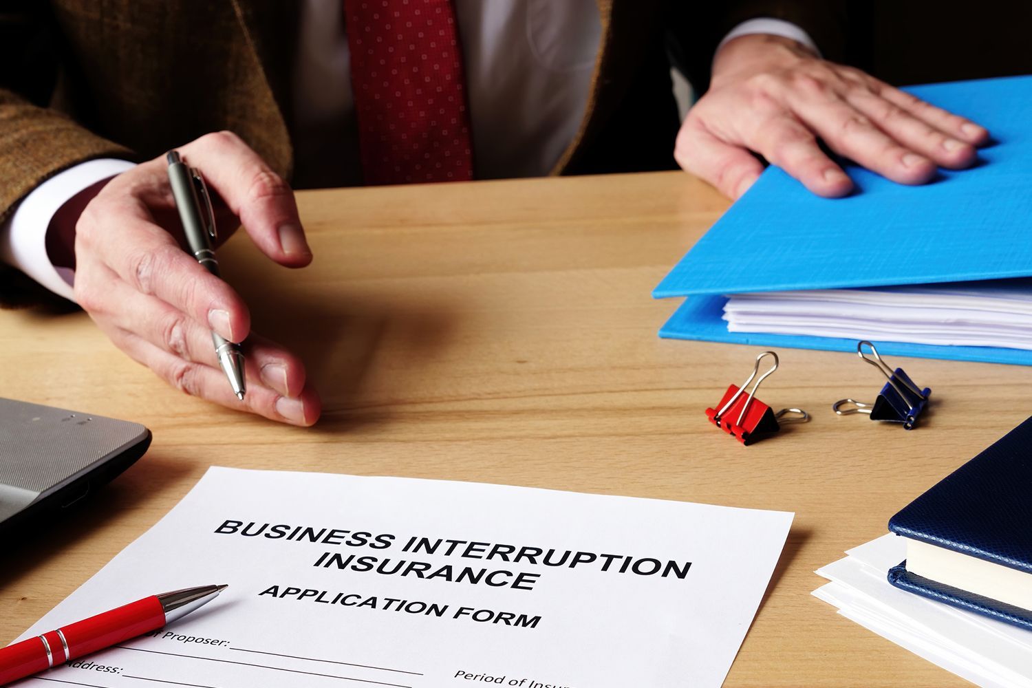Person with pen over business interruption insurance form on a desk. Blue folder, red pen, and paper clips present.