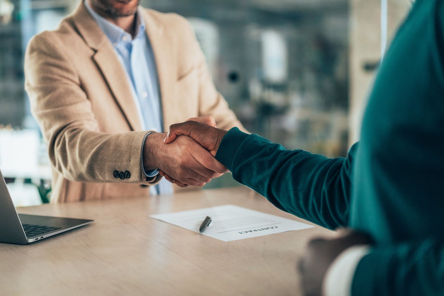 Two people shaking hands over a contract on a table. One in a tan jacket, the other in a green top.