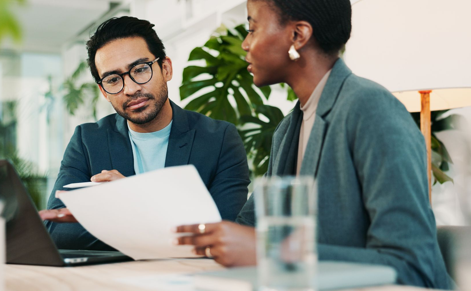 Two people in business attire at a table; one signing a document, another holding it.