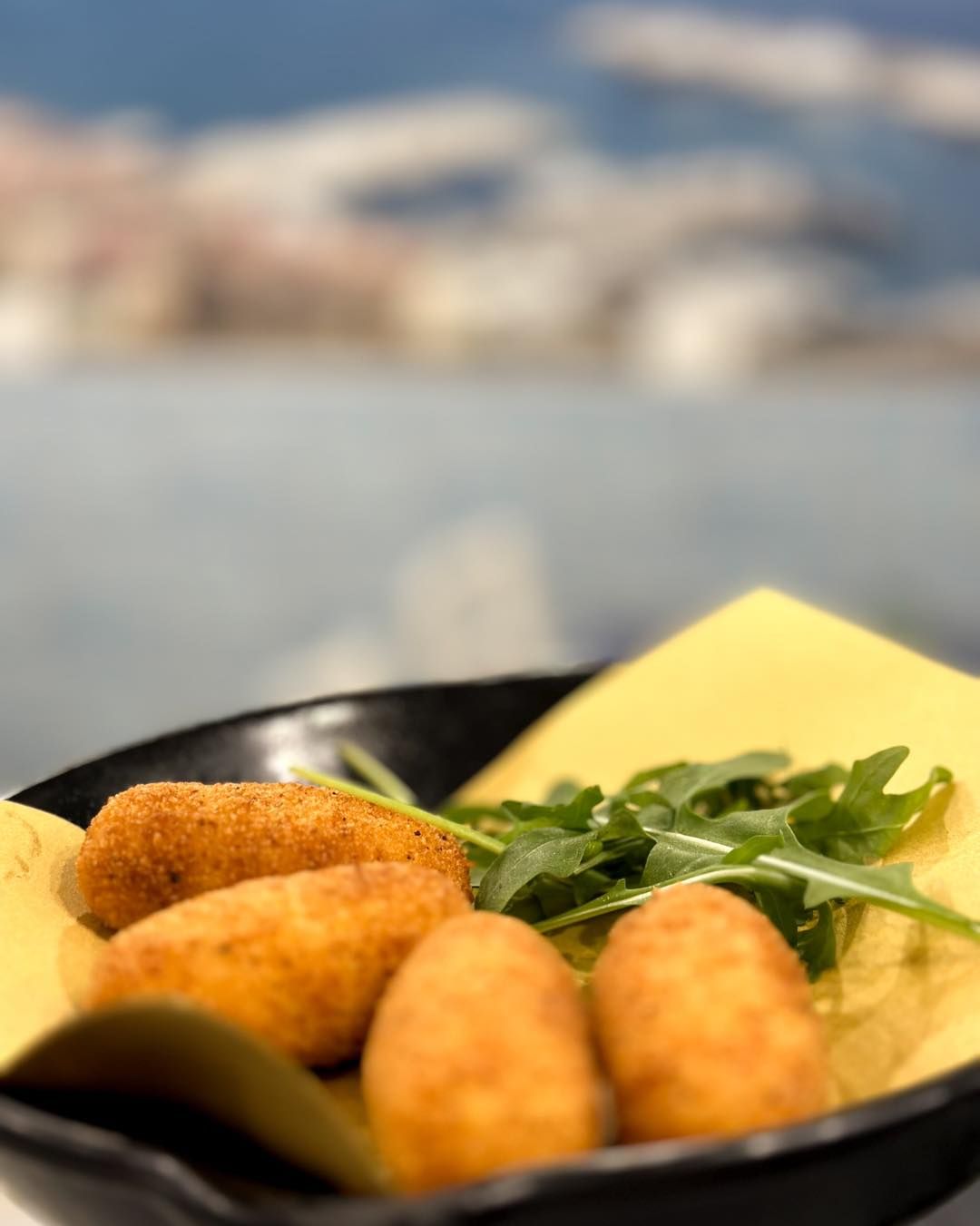 Crocchette fritte con verdure su carta gialla in un piatto nero, sfondo sfocato.