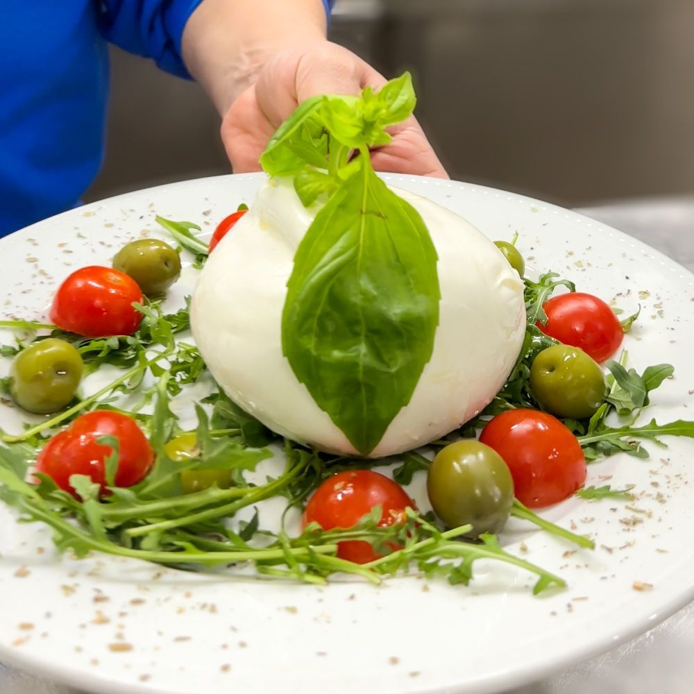 Burrata con basilico, pomodori, rucola e olive su un piatto bianco.