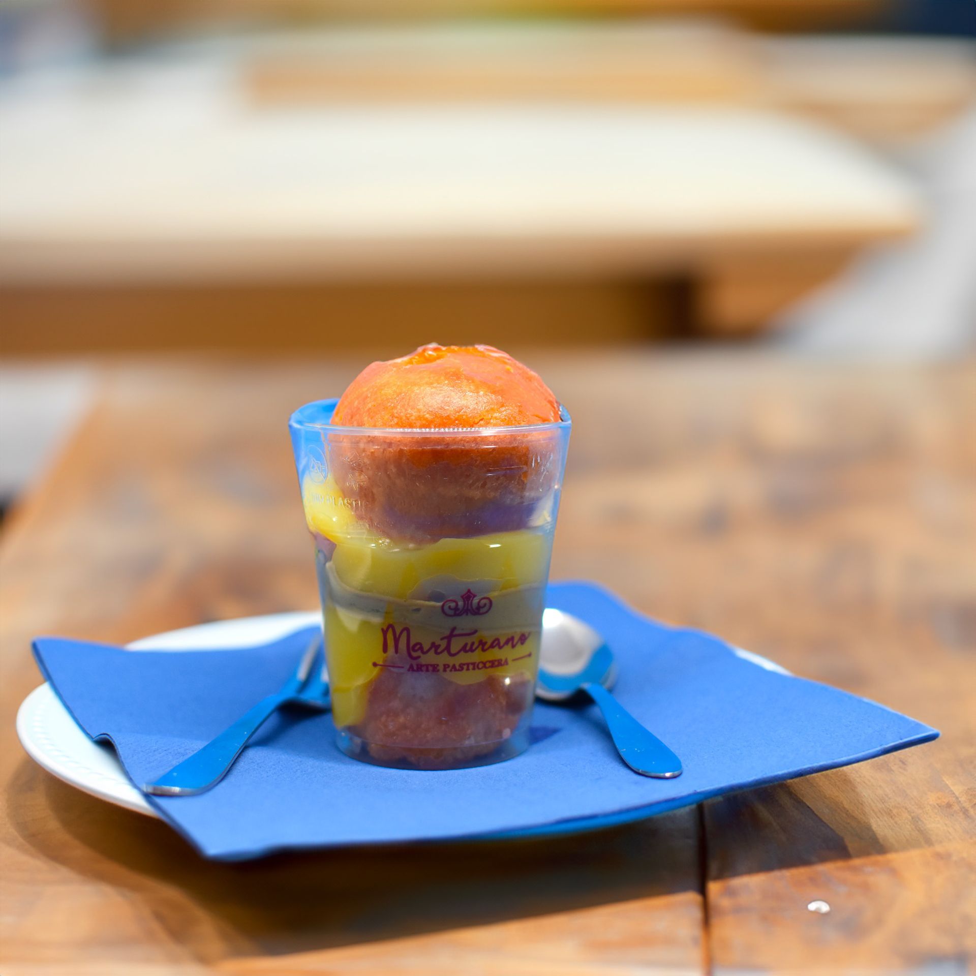 Dessert servito in una tazza trasparente con tovagliolo blu e cucchiai su un piatto bianco appoggiato su una superficie di legno.