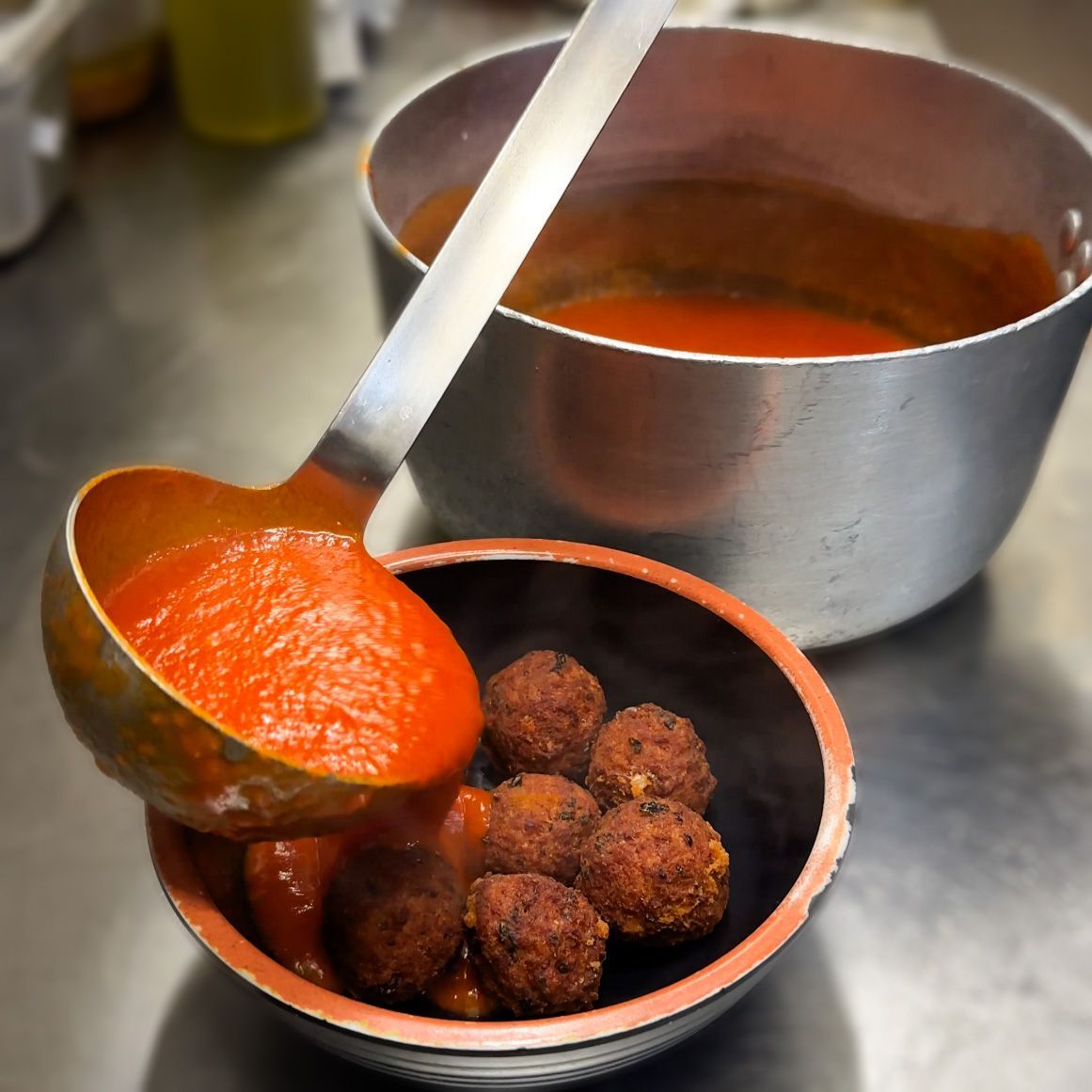 Un mestolo versa la salsa di pomodoro sulle polpette in una ciotola di terracotta; sullo sfondo, una pentola di salsa.