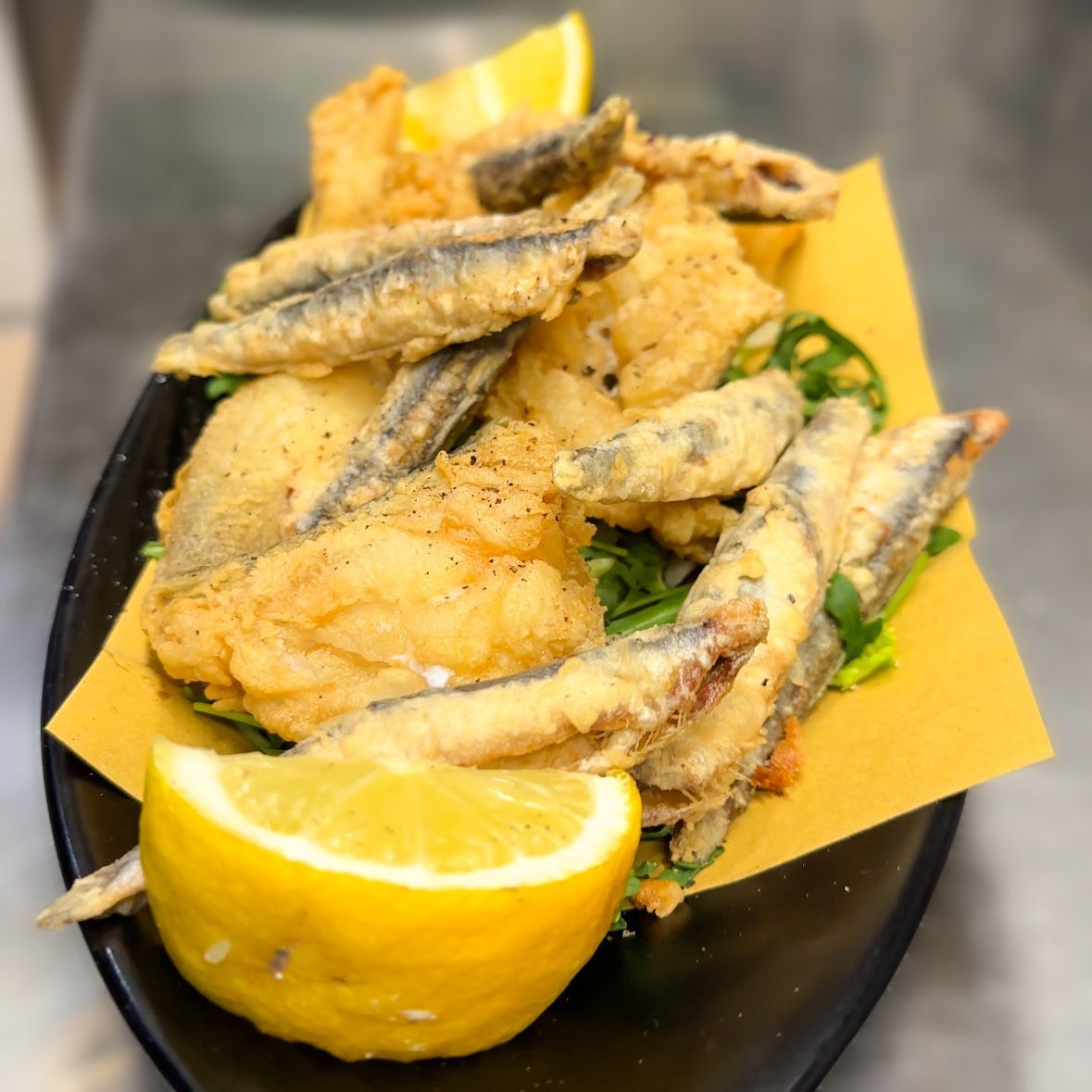 Pesce fritto e spicchi di limone su un piatto nero con carta da forno gialla e guarnizione verde.