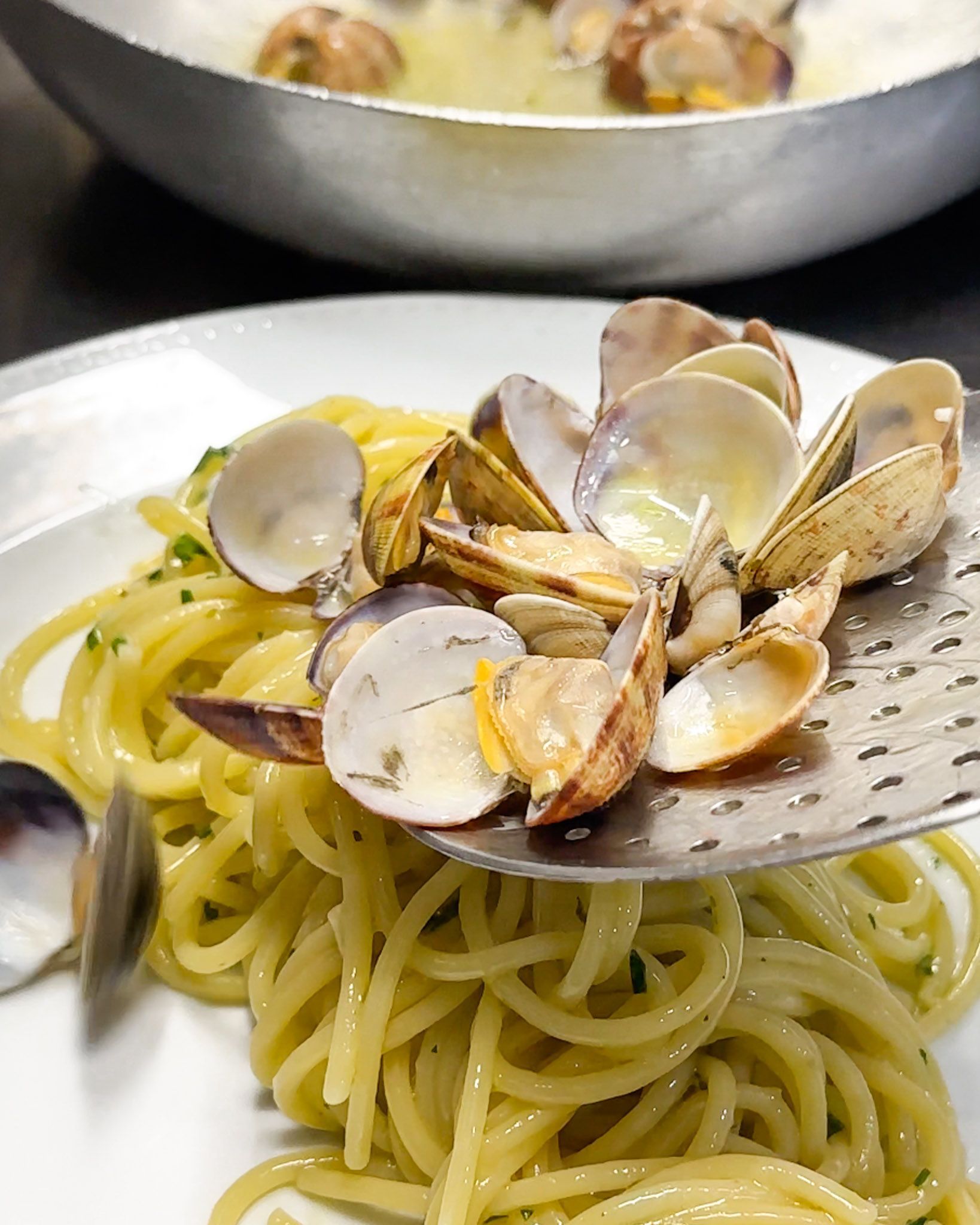 Spaghetti alle vongole aggiunti a un piatto con una schiumarola; sullo sfondo è visibile una pentola di metallo.