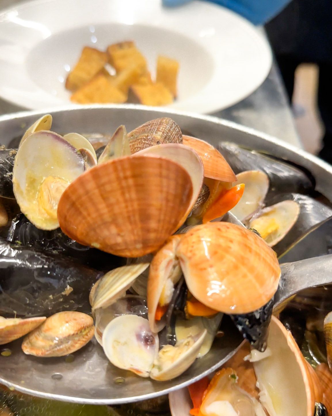 Primo piano di vongole e cozze in una ciotola di metallo, con un piatto separato di crostini sullo sfondo.