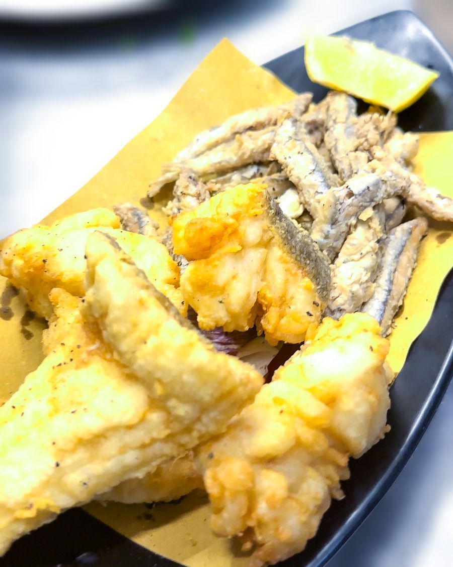 Piatto di pesce fritto con spicchio di limone su carta gialla e piatto nero.
