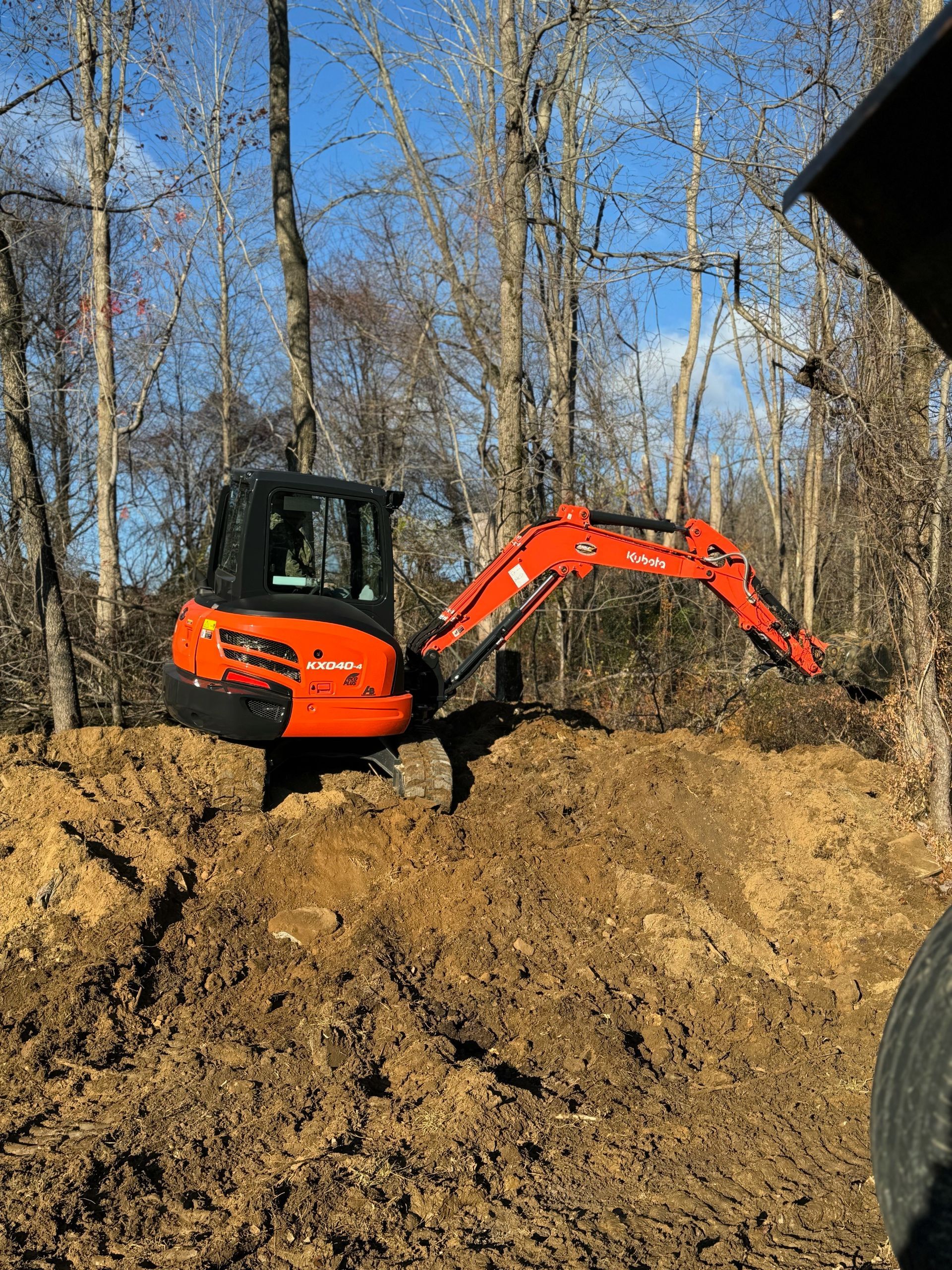 A small orange excavator is digging in the dirt in the woods.