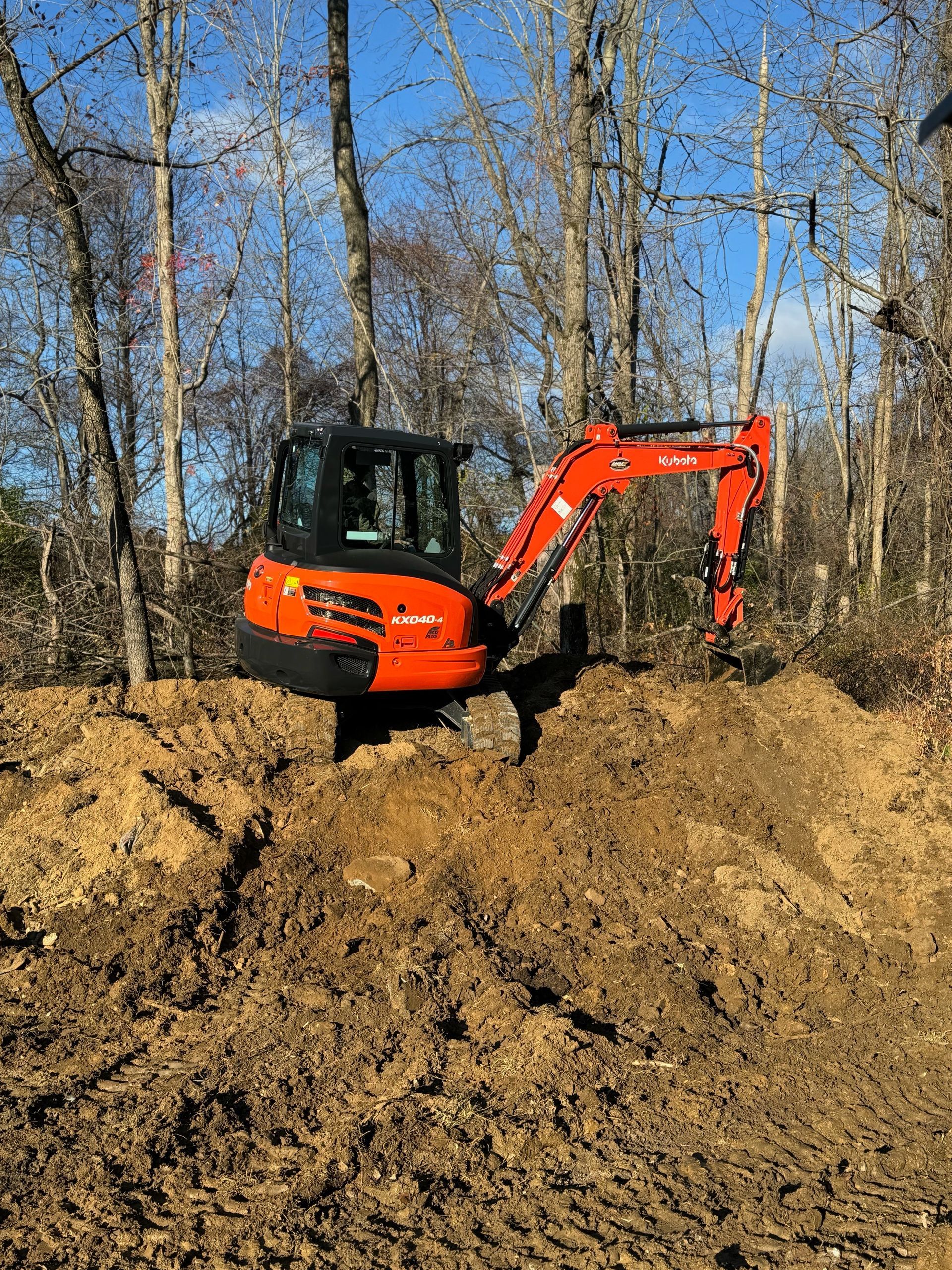 An orange excavator is digging a hole in the dirt in the middle of a forest.