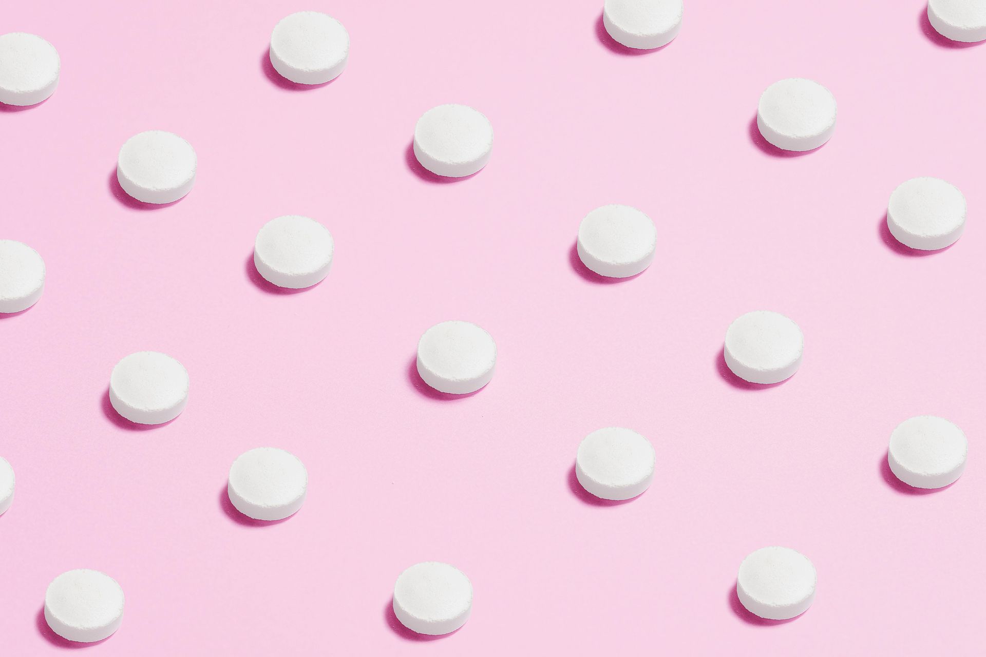 White pills arranged on a pink background, evenly spaced, casting shadows.