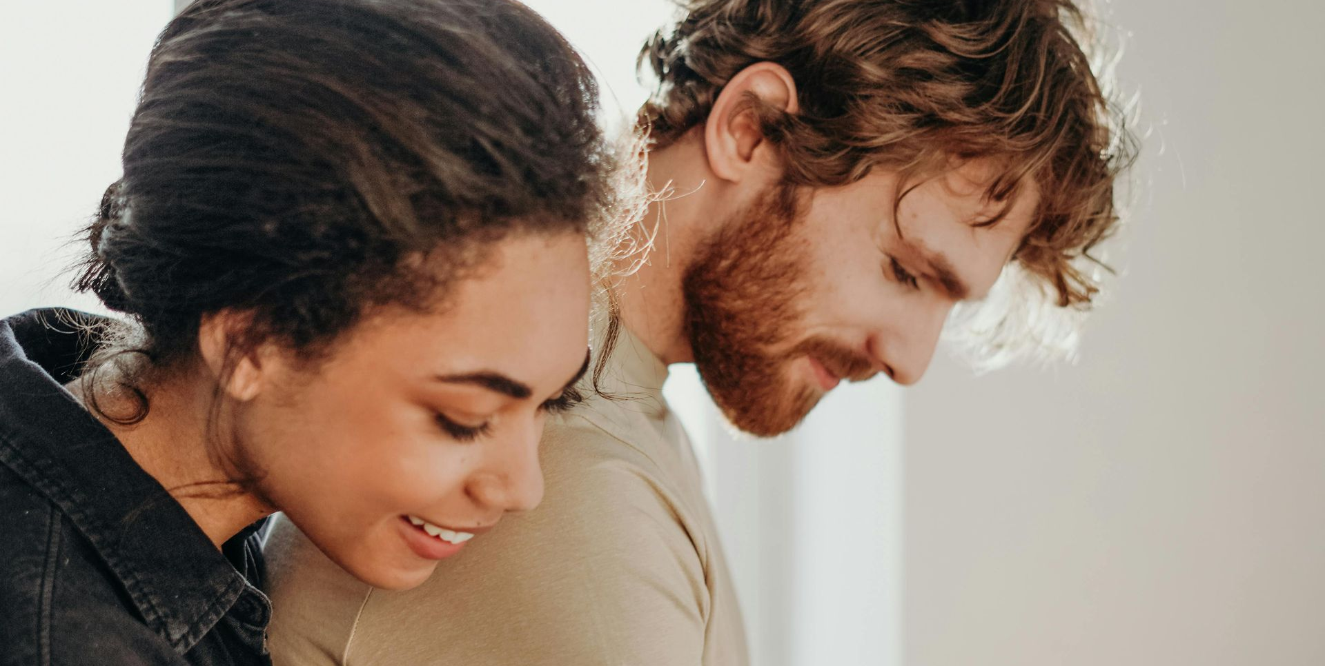 Couple looking down, smiling. Woman with dark hair, denim jacket. Man with red beard, light shirt, neutral background.