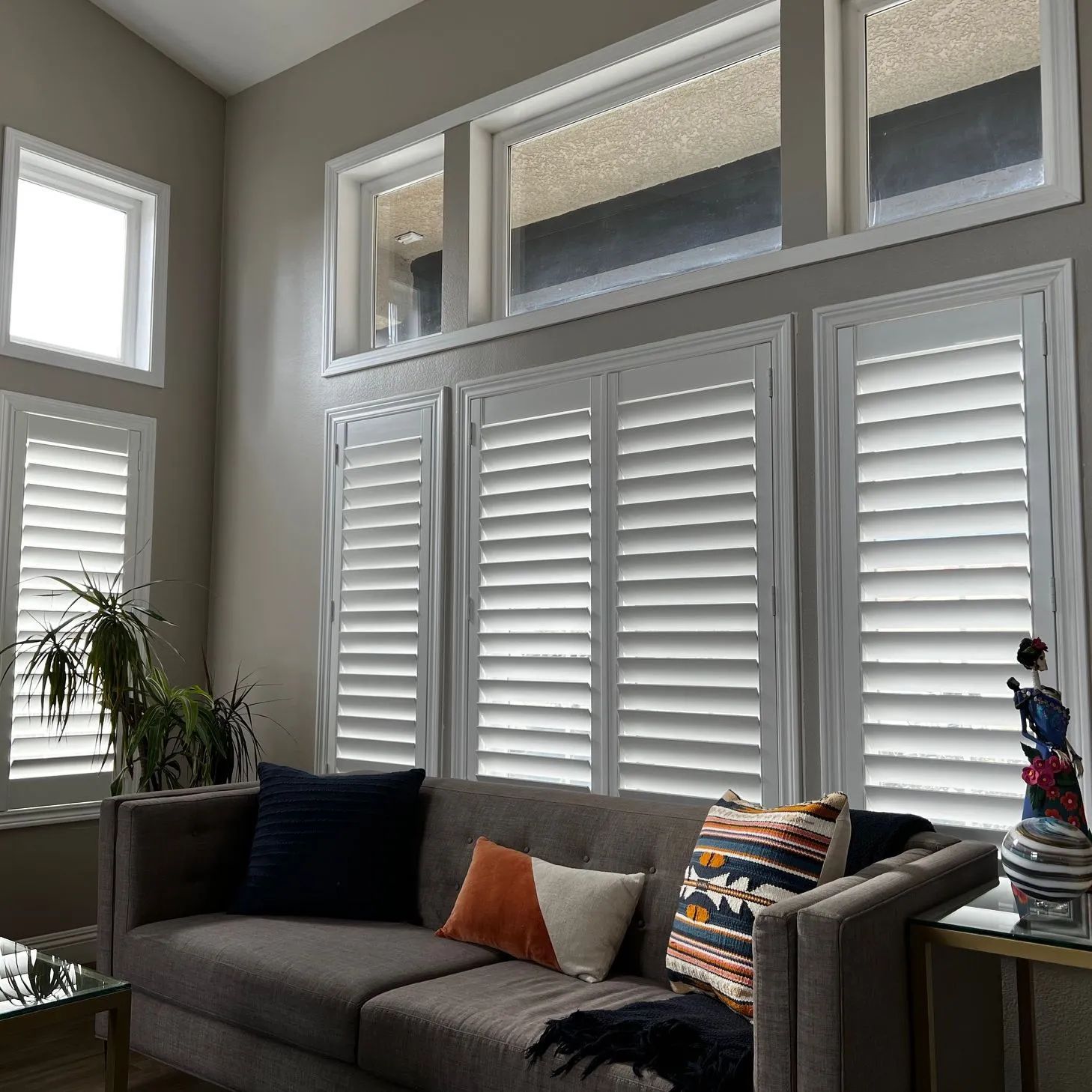 A gray sofa sits in front of tall windows with white plantation shutters in a brightly lit, neutrally toned living room.