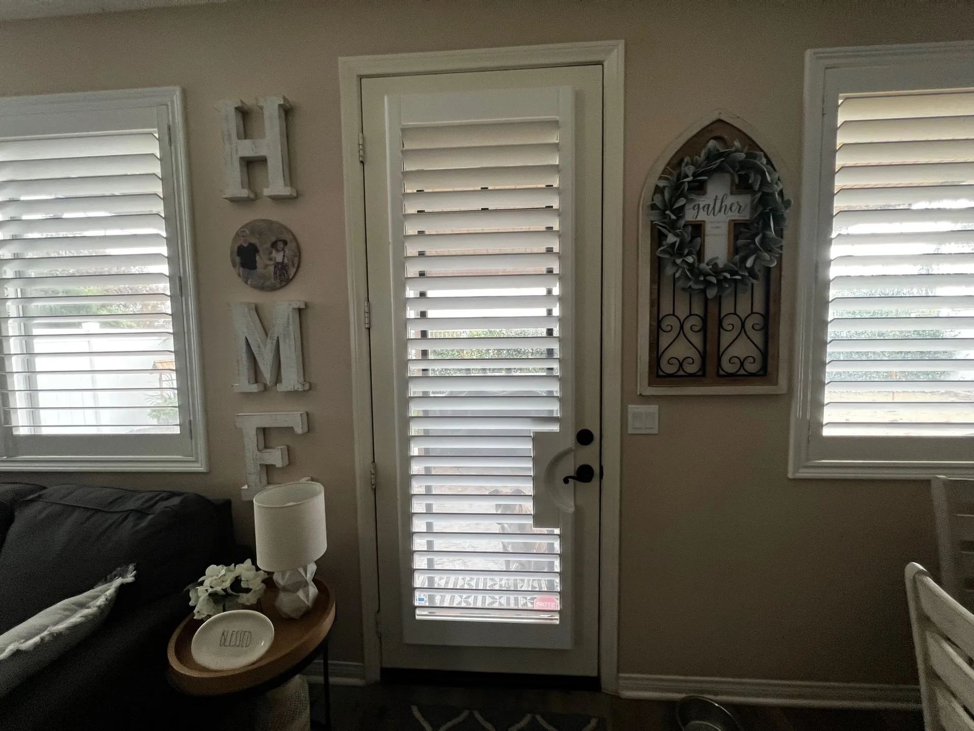 A room interior featuring a white door with plantation shutters, flanked by two windows, home decor, and a side table.