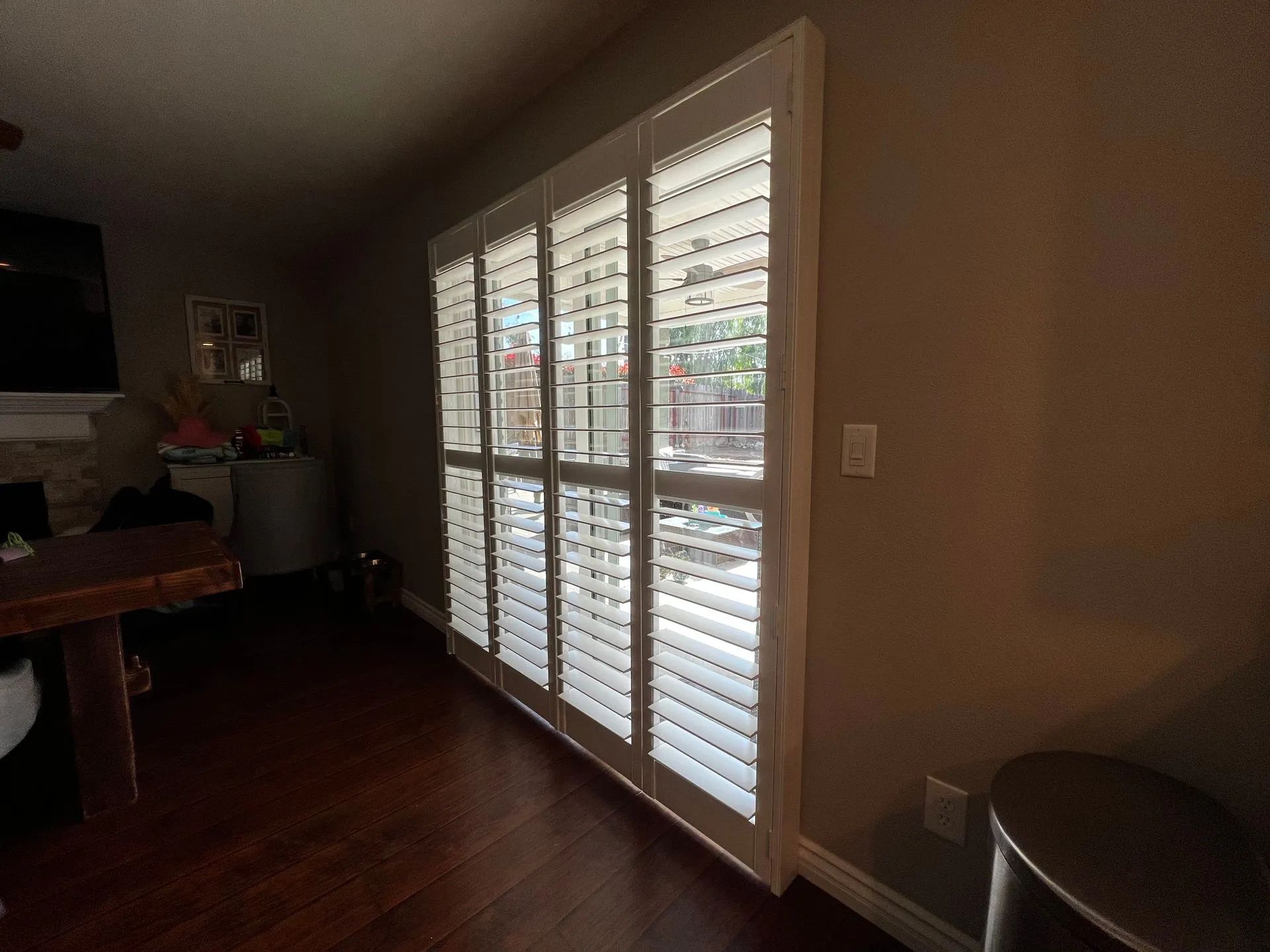 Indoor view of window shutters. Sunlight streams through them into a room with dark hardwood floors and beige walls.