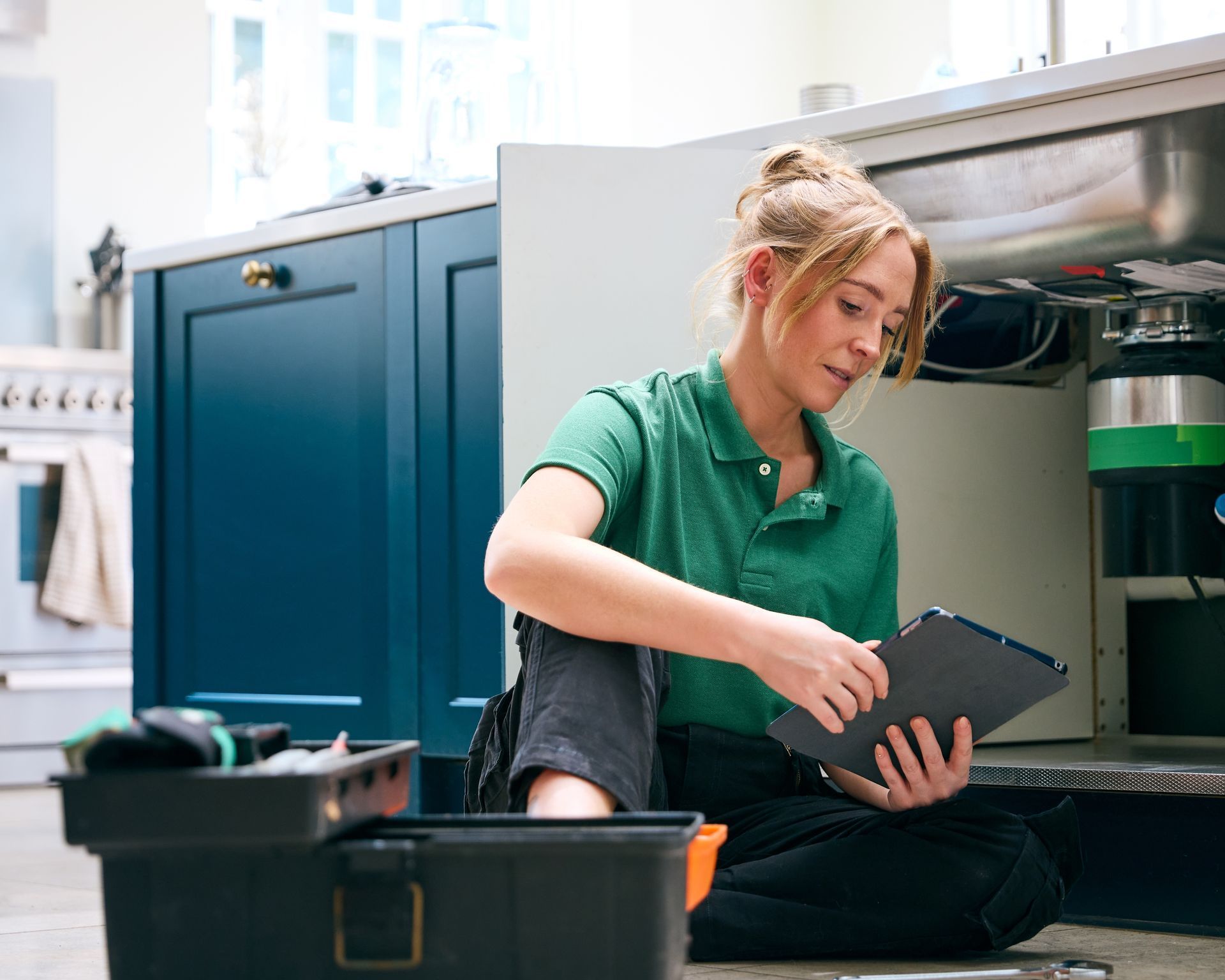 A woman is sitting on the floor in a kitchen looking under a sink.