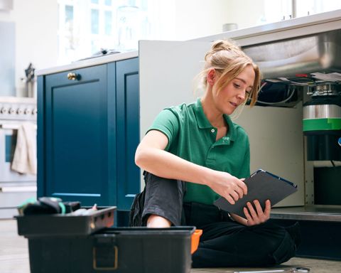 A woman is sitting on the floor in a kitchen looking at a clipboard.