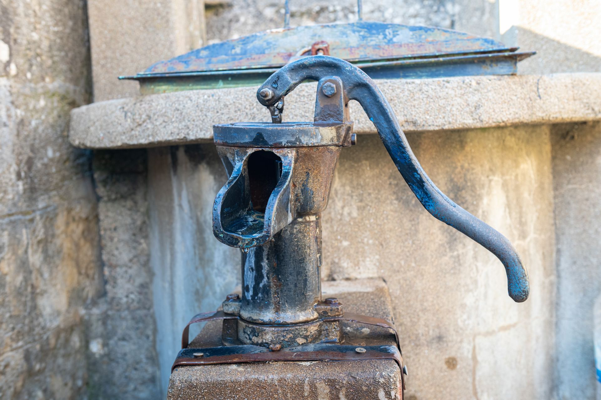 An antique black cast-iron hand water pump mounted on a stone base in front of a concrete well structure. An antique black cast-iron hand water pump mounted on a stone base in front of a concrete well structure.