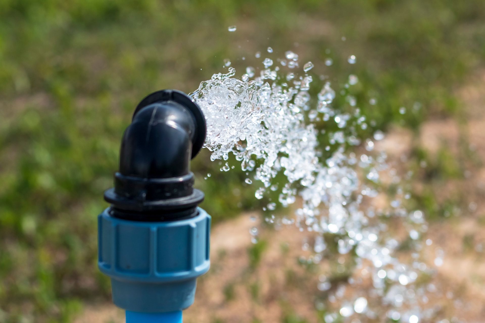 Water sprays from a black angled pipe fitting with a blue connector against a blurry green and brown natural background. Water sprays from a black angled pipe fitting with a blue connector against a blurry green and brown natural background.